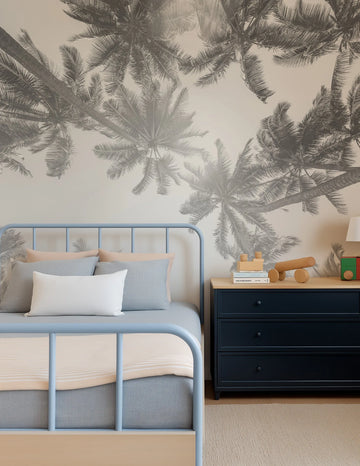 "Peel and stick wallpaper mural featuring an overhead view of palm trees in a monochrome grey and white palette, creating a serene, tropical atmosphere in a child's bedroom. The wallpaper is displayed behind a light blue metal bed frame with a pale blue duvet and pillows, and a white rectangular pillow. A dark blue dresser with a light wood top sits next to the bed, adorned with wooden toys and books, and a white lamp."
