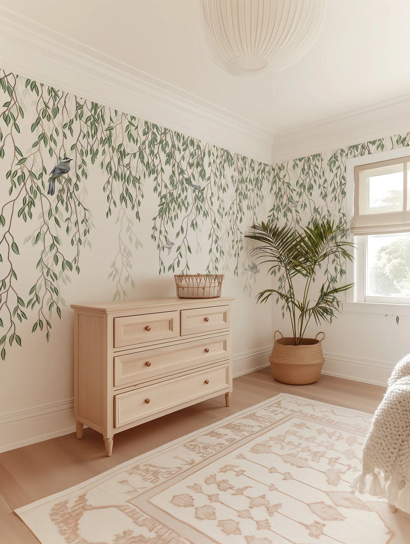 Peel and stick wallpaper featuring a mural of delicate willow branches with small green leaves cascading downwards on a warm cream background, with several small grey birds perched among the branches. The wallpaper is displayed on two walls of a bright bedroom, above a light wood dresser with brass knobs and beside a window with a cream roman blind. A large green palm plant in a woven basket sits in the corner, and a cream patterned rug covers the light wood floor.