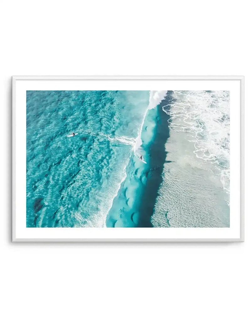 Framed aerial photograph of a coastal beach scene with a white frame and white border. The composition features two surfers, one paddling on a board in the turquoise ocean and another standing on a surfboard riding a white wave towards the sandy shore. The clear water reveals the textured seabed.