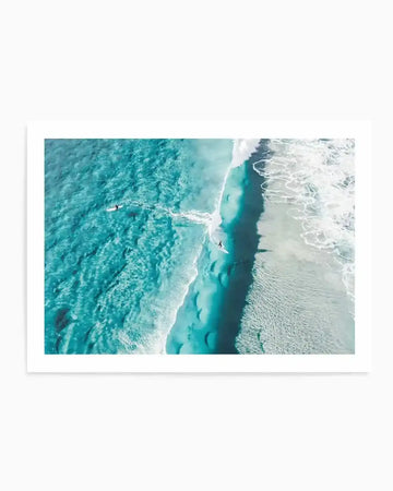 Aerial photograph art print of two surfers in turquoise ocean water near Mornington Victoria, with white foaming waves and a dramatic dark underwater trench visible from above. The image captures the contrast between shallow turquoise waters on the left, a deep navy channel in the center, and white-capped waves on the right shore. Printed on white paper with white border and displayed in a white frame.