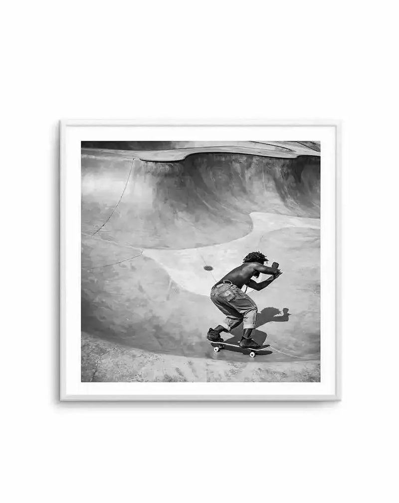 Framed black and white photograph of a skateboarder in a concrete skate park, with a white frame and white border. The composition features a shirtless man with dreadlocks, wearing loose-fitting pants, riding a skateboard on a curved ramp, captured mid-action with his shadow cast behind him.