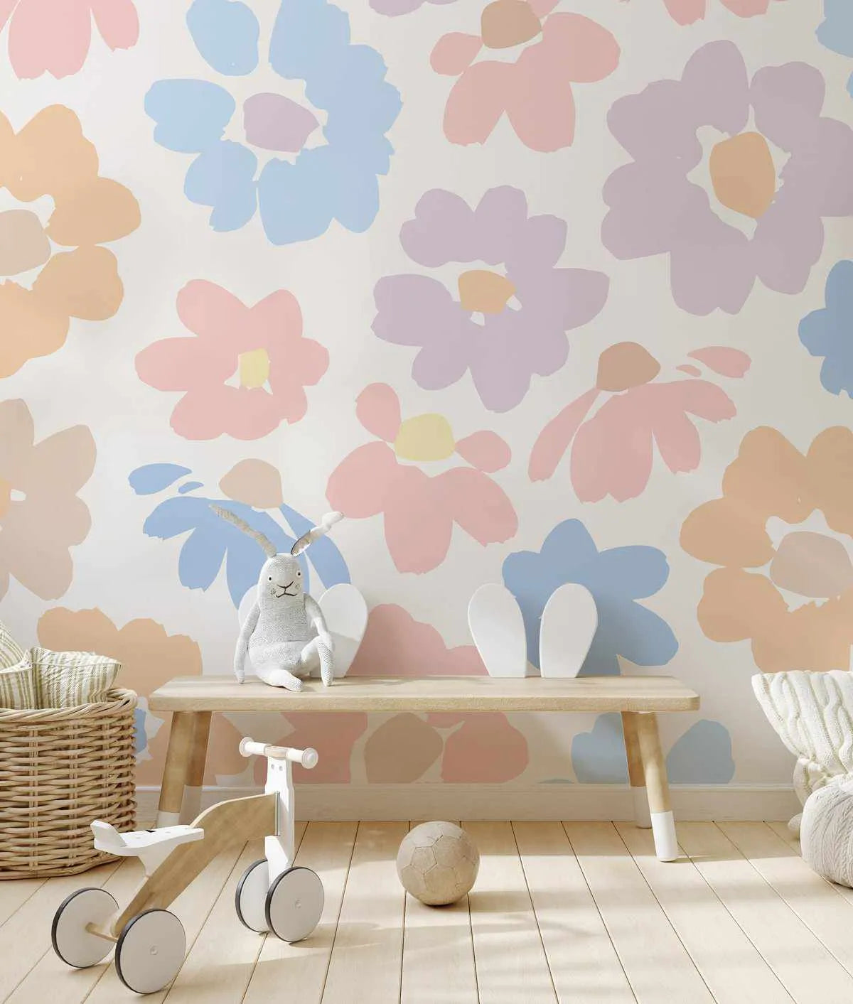 Nursery:
Peel and stick wallpaper featuring a repeating pattern of large, hand-painted flowers in soft pastel shades of peach, baby blue, light pink, and lavender on a white background, displayed in a child's room. The room includes a light wood bench with a stuffed bunny and white bunny ear decor, a woven basket, a wooden balance bike, and a textured ball on a light wood floor.