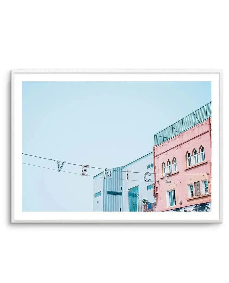 Framed photography art print of the iconic Venice Beach sign, featuring the letters "VENICE" strung across a light blue sky, with a white building and a salmon pink building with arched windows visible below. The print is displayed in a white frame with a white border.