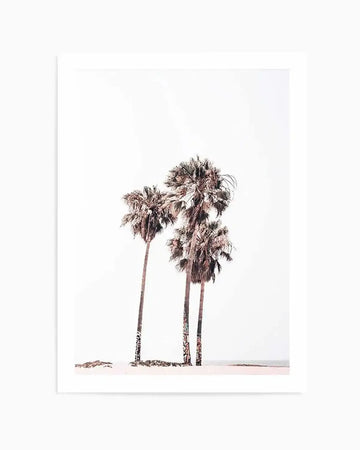 "Framed photography art print featuring three tall palm trees on a sandy beach with a light, almost white sky and subtle ocean horizon. The palm tree trunks have visible dark markings, possibly graffiti, and the overall image has a soft, muted, and slightly desaturated colour palette."