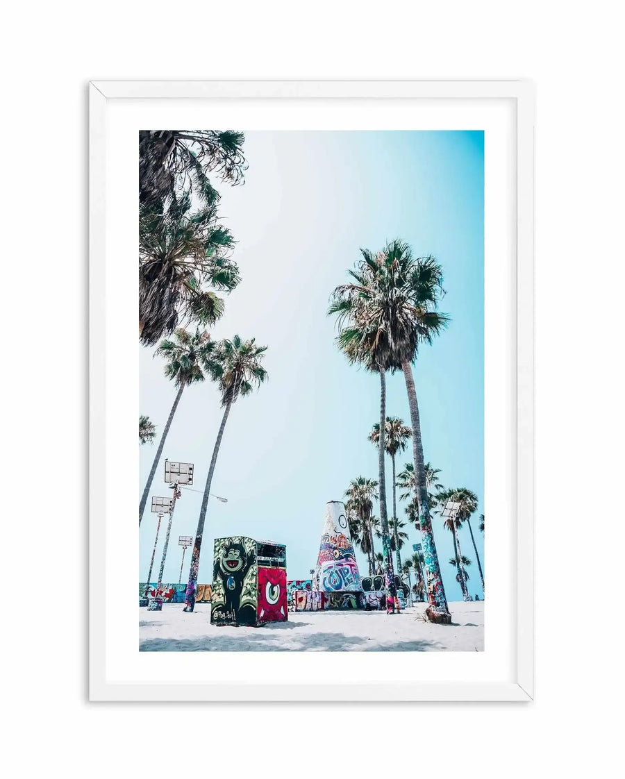 Framed art print featuring a photograph of Venice Skate Park with tall palm trees against a clear turquoise sky. The sandy beach below displays colorful graffiti-covered skate ramps and structures in red, black, white, and pink tones. Street lights and additional palm trees line the scene. The photograph is mounted in a white frame with a white border.