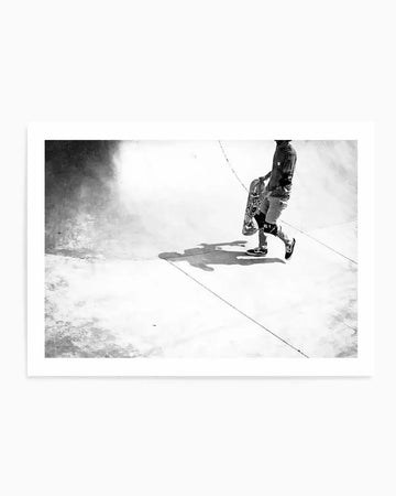 Framed black and white photograph of a skateboarder walking through a skate park, with a natural wood frame and white border. The composition features a person in dark clothing and knee pads carrying a skateboard, casting a long shadow on the light-colored concrete ground. The background shows the curved walls of the skate park.