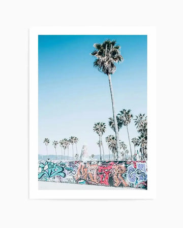 "Framed photography art print capturing a vibrant outdoor scene with a clear blue sky and numerous palm trees. The foreground features a concrete wall covered in colourful graffiti in shades of teal, orange, red, purple, and black. A tall palm tree stands prominently in the center-right, reaching towards the sky, while other palm trees are visible behind the graffiti wall. The overall mood is bright and energetic, suggestive of a sunny, urban beach environment."