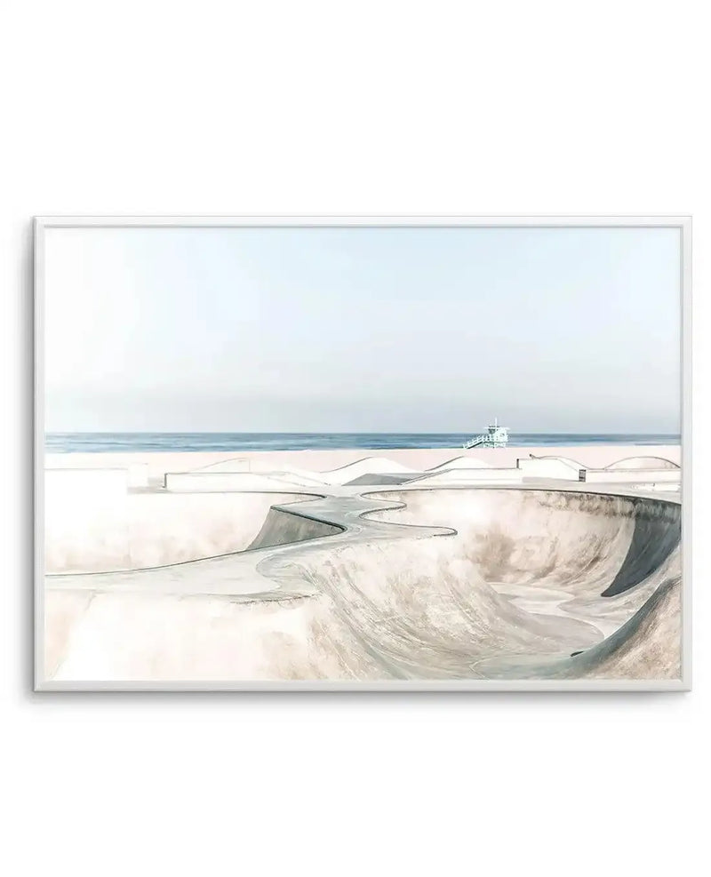 Framed art print depicting an architectural illustration of a concrete skate park with curved ramps and bowls in muted grays and taupes. The park overlooks a calm sea and sandy beach under a pale blue-gray sky. A lifeguard tower sits on the distant shoreline. The watercolor-style illustration has soft, blended tones with architectural detail in the foreground concrete structures. The print is displayed in a silver-toned frame with white border.