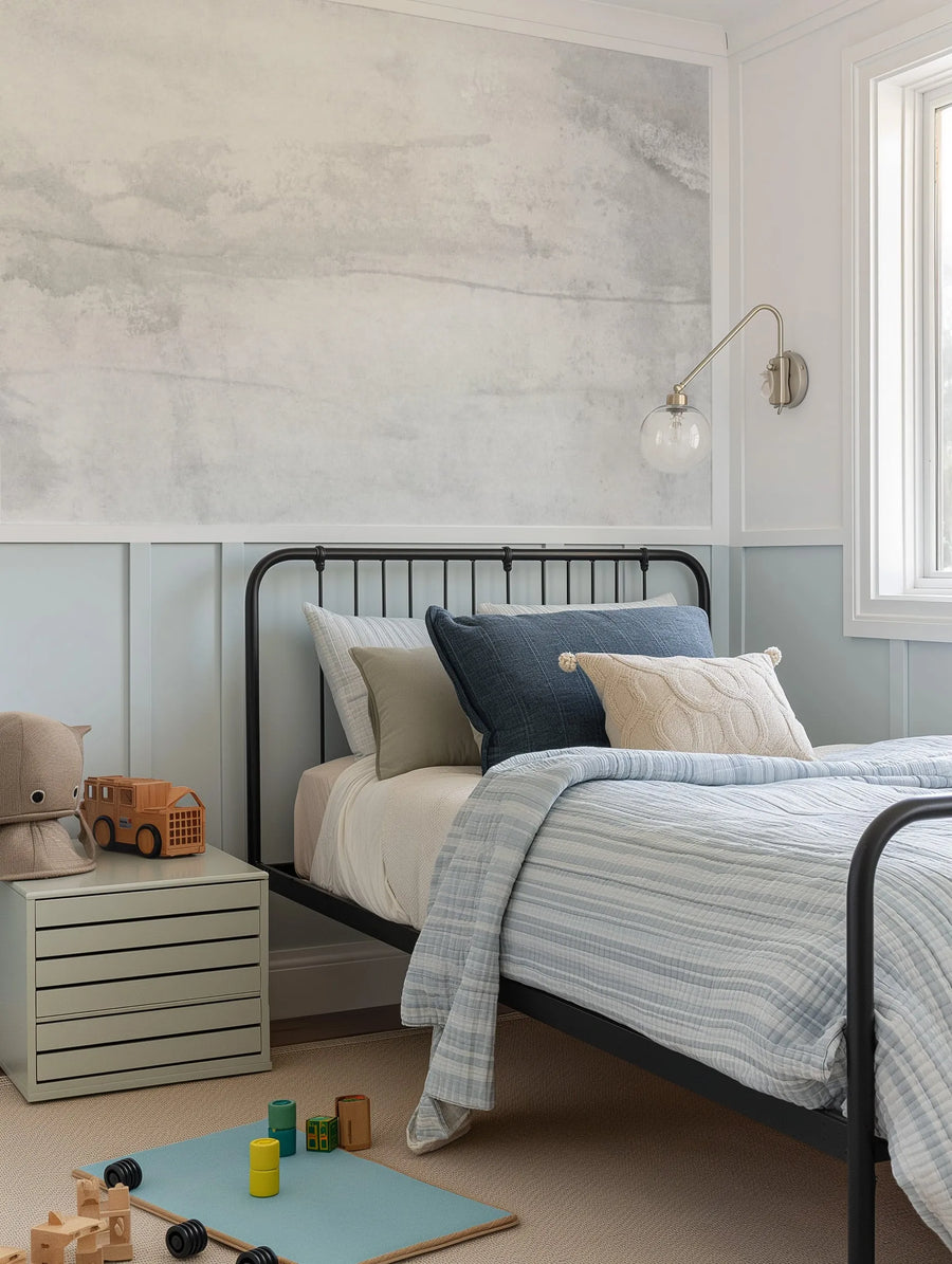 Lifestyle shot of a child's bedroom featuring Vienna Grey Wallpaper above a light blue wainscoting. The wallpaper has a soft, mottled grey and white pattern resembling a cloudy sky or subtle marble texture. A black metal bed frame with blue and cream bedding is centered in the room, with a light grey nightstand and wooden toys on the floor.