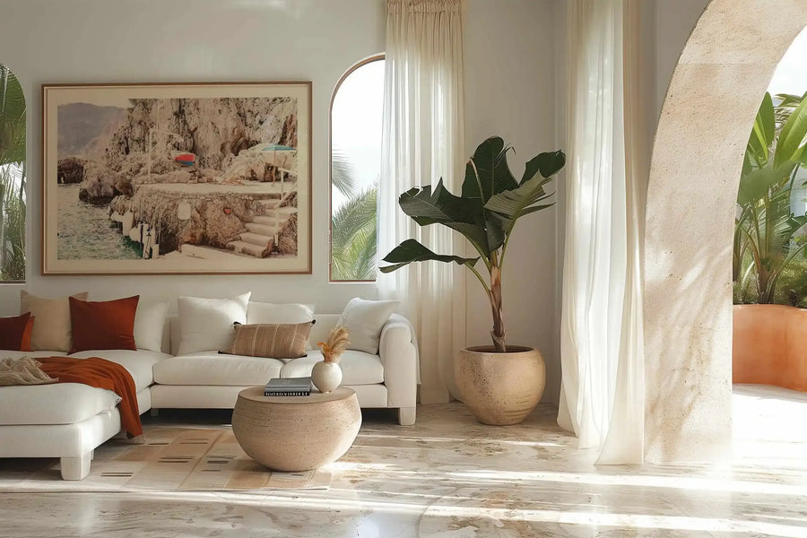 Lifestyle shot of a minimalist living room featuring a large framed art print displayed above a white sectional sofa. The print is a photograph of a rocky coastal scene with a small beach and steps leading down to the water, with a red and blue umbrella visible on the rocks. The art print has a natural wood frame and a white border. The room includes a round stone coffee table, a large potted banana plant, and an arched window with sheer white curtains.