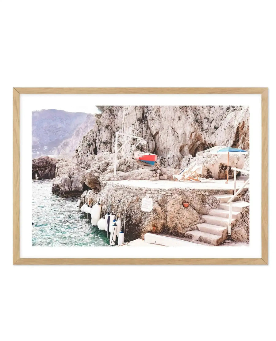 Framed photography art print of a coastal scene with a natural wood frame and white border. The image captures a rocky coastline with clear turquoise water, a small red and blue boat suspended above a stone platform, and a set of stone stairs leading down to the water. Two white beach chairs and a blue and white striped umbrella are visible on the platform, along with several white buoys floating in the water. The mood is bright and serene.