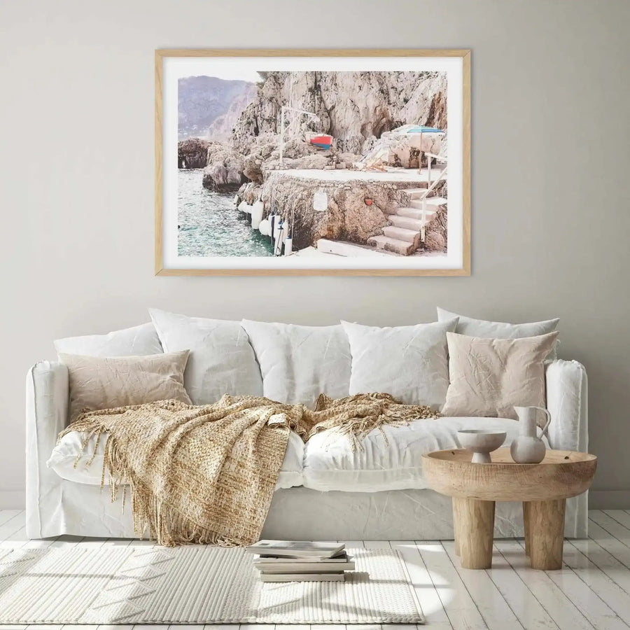 Lifestyle shot of a minimalist living room featuring a framed photography art print above a white slipcover sofa. The print depicts a rocky coastline with turquoise water, a small red and blue boat, and steps leading down to the water, framed in natural wood with a white border. The sofa is adorned with cream pillows and a textured beige throw, with a wooden side table and books on a rug completing the scene.