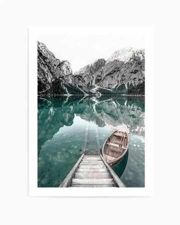 Framed black and white photograph of a mountain lake scene with a white border. The composition features towering, rugged mountains with snow-capped peaks reflecting in the calm, teal-hued water. A wooden boathouse dock extends into the foreground, with a classic wooden rowboat gently floating beside it.
