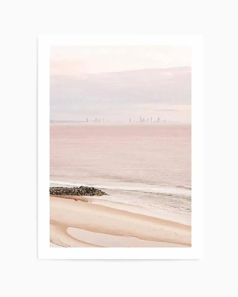 Framed photograph of a coastal beach scene with a soft, ethereal quality, featuring a wide expanse of pale pink ocean meeting a sandy beach with gentle waves. In the distance, a faint skyline of buildings is visible under a hazy, light pink sky. A small rock groyne extends into the water on the left, with a tiny red object on the sand nearby. The overall mood is serene and dreamlike.