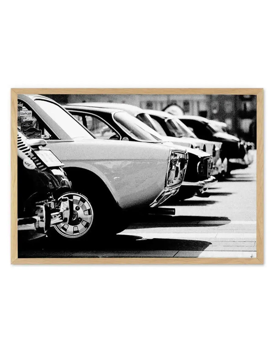 Black and white vintage photograph showing a row of classic cars parked in a line, photographed from a low angle emphasizing their sleek, curved body designs and chrome details. The composition captures the distinctive styling of mid-century automobiles with their rounded forms and decorative wheel covers. The photograph is displayed in a natural wood frame with a clean, light finish, presented as a framed art print against a plain background.