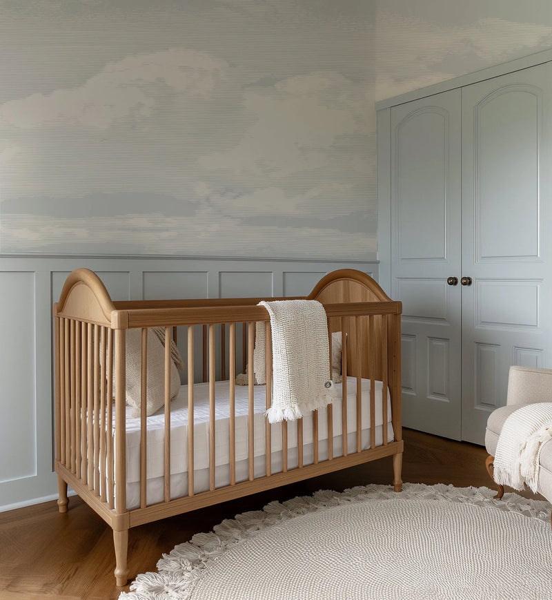 Lifestyle shot of a nursery featuring a peel and stick wallpaper mural with a vintage cloud design in soft grey and off-white tones. The wallpaper covers the upper half of the wall, above light grey wainscoting. A natural wood crib with a white mattress and a cream textured blanket is in the foreground, and a light grey closet with arched paneling is visible on the right.