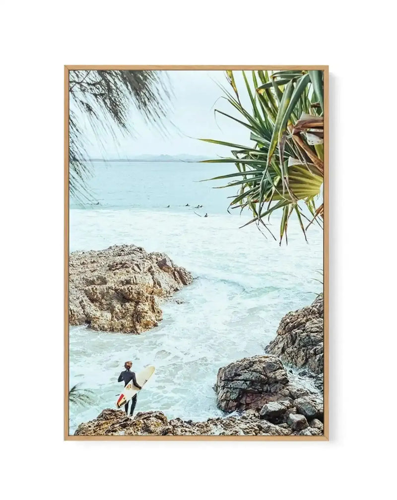 Framed canvas art print featuring a vintage-style photograph of a surfer in a dark wetsuit carrying a cream-colored surfboard, standing on rocky terrain overlooking turquoise ocean waters. Palm fronds frame the top right corner, with limestone rocks in the foreground and middle ground. Distant surfers can be seen in the calm water. The scene has soft, hazy tones with pale blue sky and translucent turquoise water typical of Byron Bay. Natural wood-tone frame with white border surrounding the artwork.