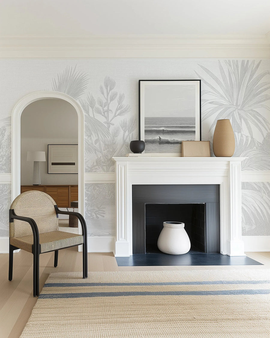 Peel and stick wallpaper mural featuring a botanical illustration in soft greige and cream tones. The design depicts layered palm fronds and tropical foliage with delicate line work, creating a serene, naturalistic pattern across the wall. Shown applied in a modern living room with a white fireplace mantel, cream upholstered chair, striped area rug, and coastal artwork, demonstrating the wallpaper's sophisticated, minimalist aesthetic against light wall surfaces.