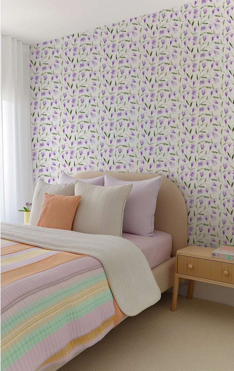 "A bedroom with a bed featuring a curved beige headboard, light pink sheets, and a striped quilt in pastel shades of pink, orange, yellow, and mint green. The wall behind the bed is covered in wallpaper featuring a repeating pattern of hand-painted violet flowers with green stems and leaves on a white background. A light wood nightstand with two drawers sits to the right of the bed, and a white curtain hangs on the left."