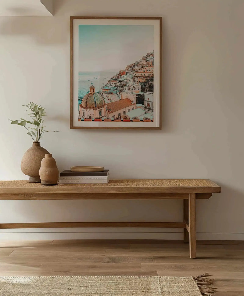 Hallway:
"Large framed art print displayed above a wooden bench in a minimalist hallway. The print features a vibrant photograph of Positano, Italy, with colourful buildings cascading down a hillside towards the turquoise sea, under a pale blue sky. The print is in a natural wood frame with a white border. On the bench, two textured terracotta vases with a green plant, a stack of books, and a wooden bowl add to the natural aesthetic."