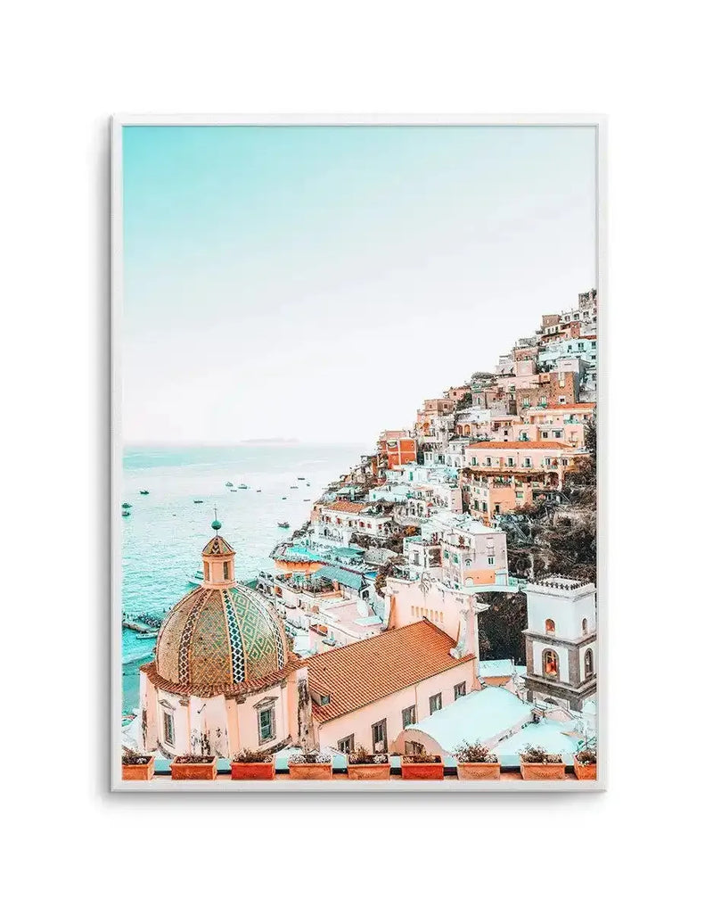"Framed photography art print capturing a vibrant, high-angle view of Positano, Italy, with a white frame. The scene features colourful buildings cascading down a steep hillside towards the turquoise Mediterranean Sea, dotted with small boats. A prominent dome with an intricate green and gold patterned roof stands in the foreground, surrounded by terracotta-roofed buildings and a distant bell tower, all under a clear, bright blue sky."