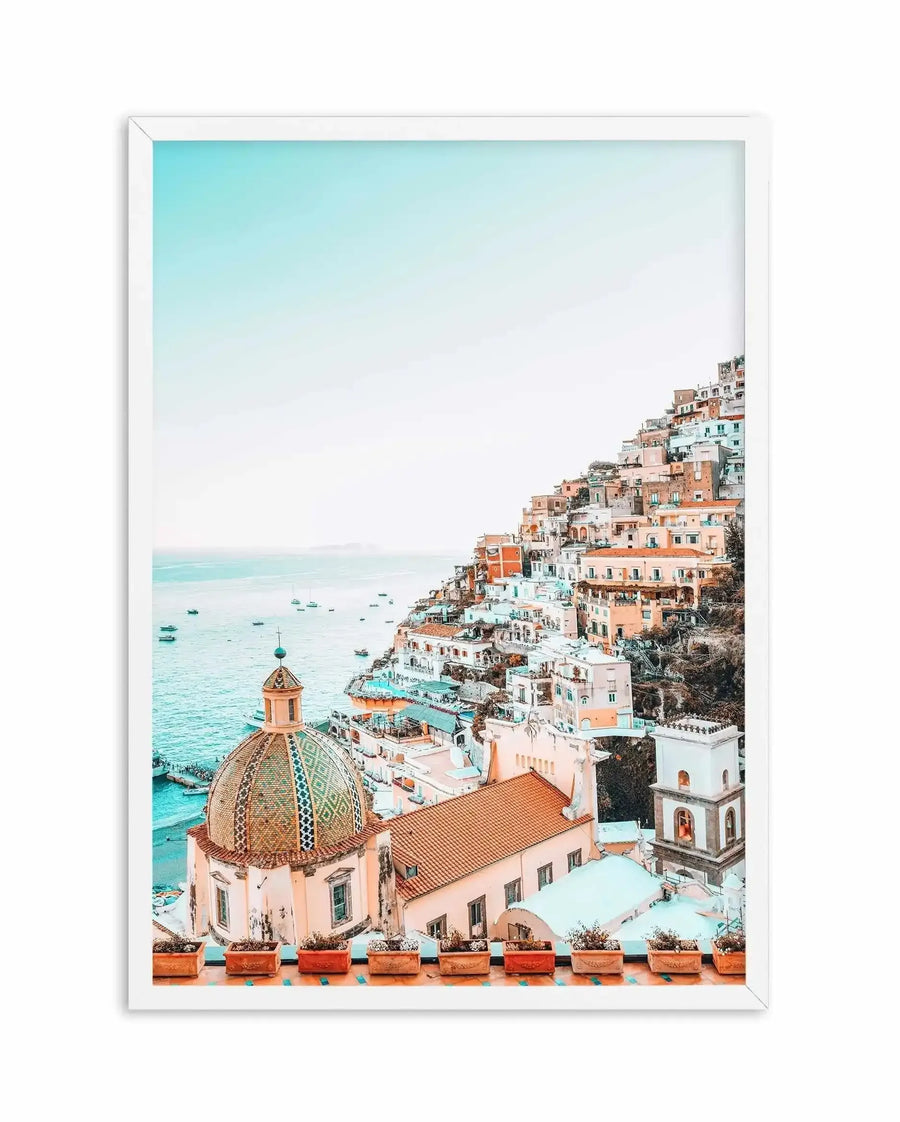 Framed photography art print of Positano, Italy, with a white frame and white border. The composition features a vibrant coastal town built into a steep hillside, with colourful buildings in shades of peach, terracotta, and white cascading down to the turquoise sea. A prominent dome with intricate green and gold tiling is visible in the foreground, and several boats dot the water. The sky is a clear, pale blue, creating a serene and picturesque mood.