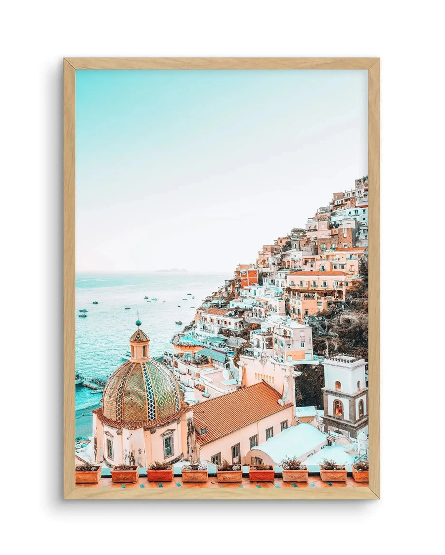 Framed photography art print of Positano, Italy, featuring a vibrant coastal town built into a hillside overlooking the turquoise sea, displayed in a natural oak frame with a white border. The composition includes colourful buildings in shades of peach, terracotta, and white, a prominent domed church with a patterned roof, and boats dotting the calm water under a bright blue sky.