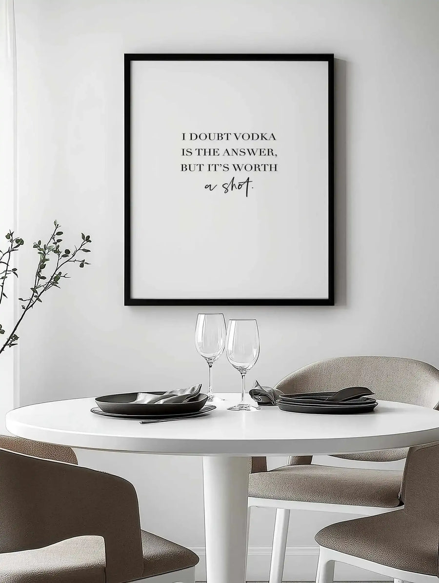 Lifestyle shot of a framed art print displayed above a white round dining table set for two. The print features the quote "I DOUBT VODKA IS THE ANSWER, BUT IT'S WORTH a shot." in black serif and script fonts on a white background, with a black frame and white border. The table is set with dark grey plates, cutlery, and two wine glasses. Two upholstered dining chairs in a light brown tone are visible.