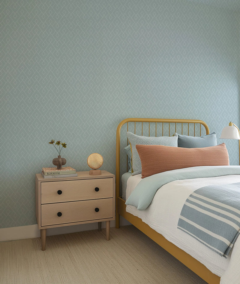 "A bedroom with light blue wallpaper featuring a repeating pattern of interlocking white diamond shapes. A mustard yellow metal bed frame is visible, dressed with a white duvet, a light blue throw blanket with white stripes, and an assortment of pillows in light blue, white, and rust orange. A light wooden nightstand with two drawers and black knobs stands beside the bed, topped with a small vase holding dried flowers and a circular woven lamp."