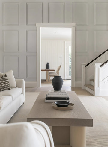 Lifestyle shot of a living room featuring wainscoting Parisian panel wallpaper in a soft grey tone. The wallpaper creates a classic, elegant backdrop with raised rectangular panels. A white sofa with a striped throw pillow is visible in the foreground, and a light wood coffee table with books and a black vase sits on a textured cream rug. Through an open doorway, a dining area with a wooden table and chairs is visible, and a white staircase with a dark wood handrail is on the right.