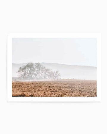 Framed photography art print depicting a misty rural landscape with a natural wood frame and white border. The composition features a field of dry, light brown grass in the foreground, leading to a line of bare trees partially obscured by a thick, pale grey mist. A faint fence line runs through the mist, and distant hills are barely visible through the haze.