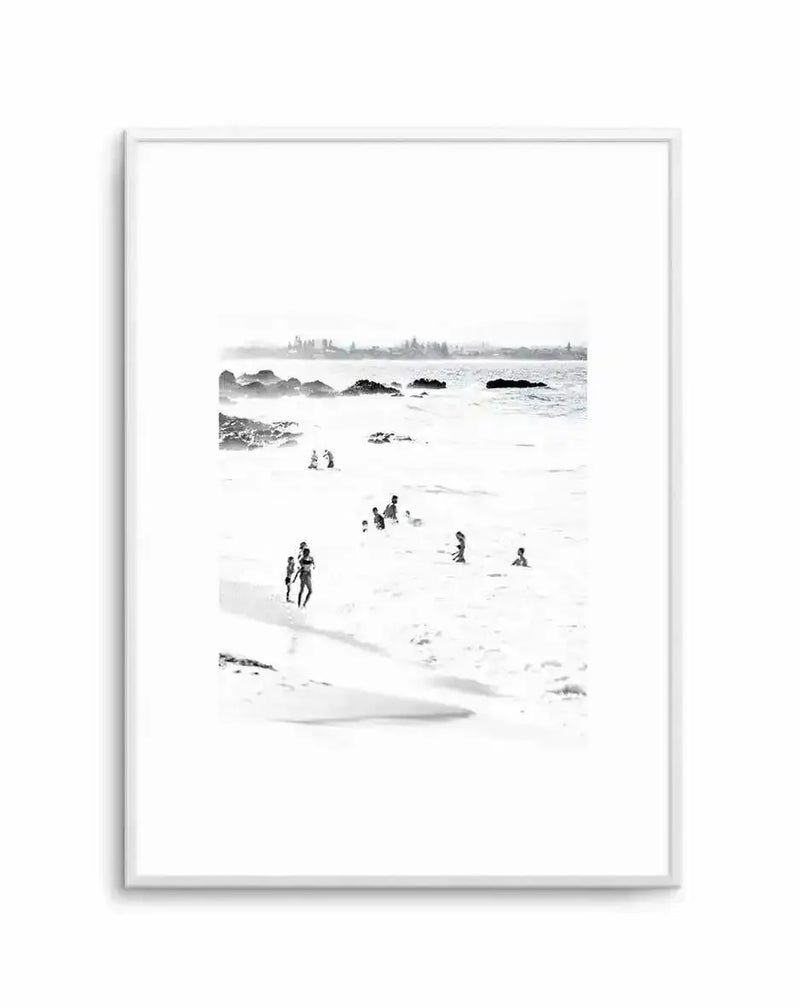 Black and white photograph titled "Wategos II" displayed in a light gray frame with white border. The minimalist coastal scene shows a sandy beach with scattered figures wading and swimming in calm water. Dark rocks and seaweed protrude from the shallow ocean. In the distance, a tree-lined shoreline and headland fade into mist. The high-contrast monochromatic print has a serene, documentary quality with soft focus creating atmospheric depth. The photograph is vertically oriented on white background.