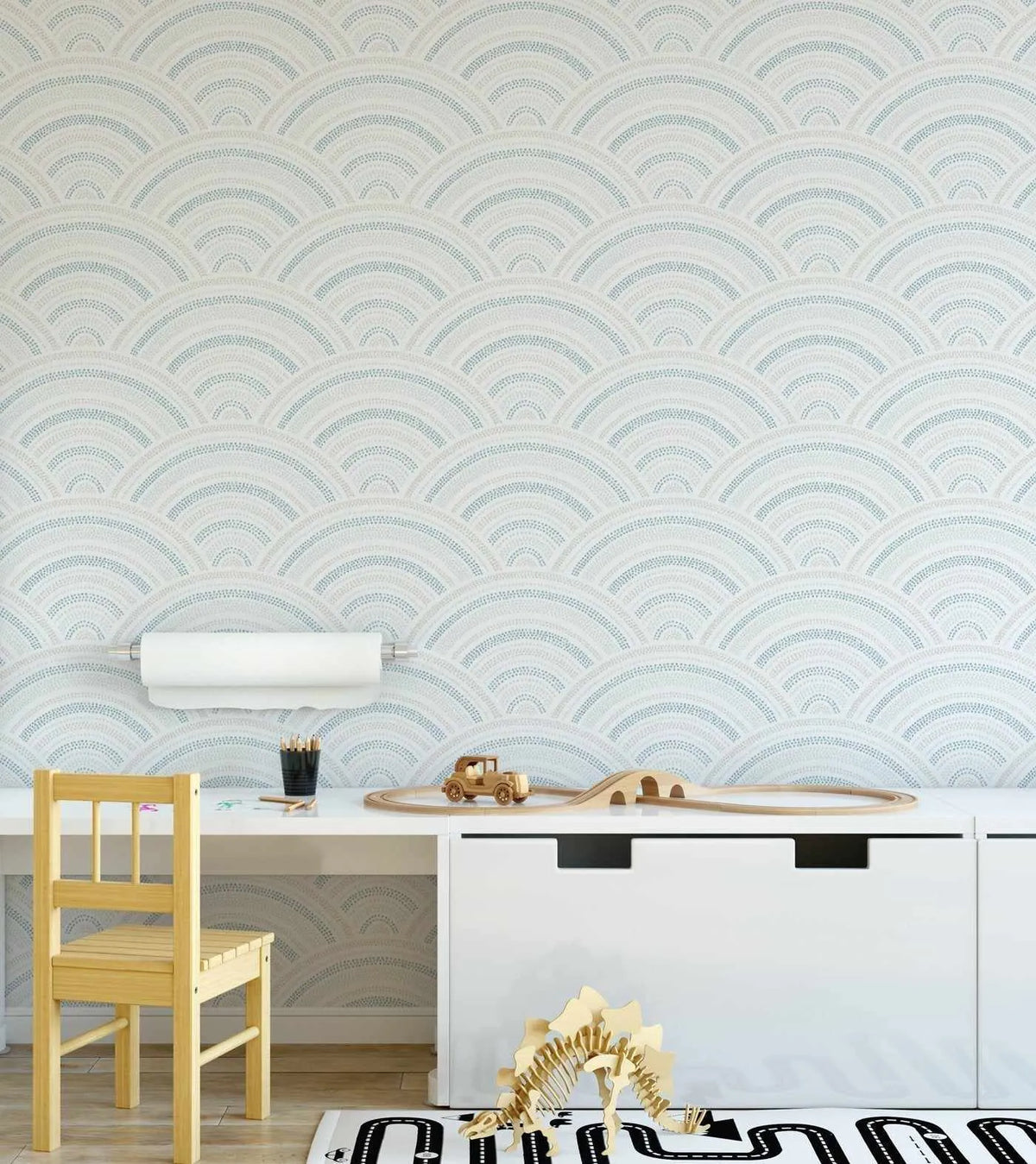 Lifestyle shot of a child's play area featuring Water Rainbows Wallpaper. The wallpaper has a repeating pattern of scalloped arches in alternating rows of light blue and pale beige dots on a white background, creating a subtle wave-like effect. A light wood child's chair is pulled up to a white desk with a paper roll holder mounted on the wall above it. On the desk are pencils, a wooden toy car, and a wooden train track. A wooden dinosaur skeleton toy sits on a road-patterned rug on the floor.
