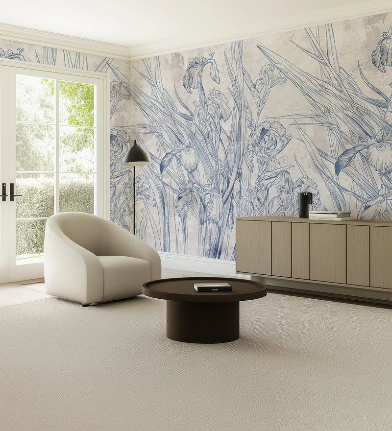 Water Iris Mural Wallpaper displayed on a feature wall in a minimalist living room. The wallpaper features a large-scale botanical illustration of water irises in deep indigo blue with fine line work and visible sketch texture, set against a distressed, textured cream background. The room includes a curved cream armchair, a dark brown round coffee table with a book, a black floor lamp, and a light wood sideboard, all on a textured cream carpet.