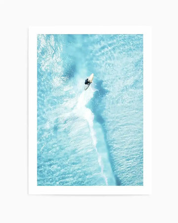 Framed aerial photograph of a surfer riding a wave in bright turquoise water, with a white frame and white border. The composition captures the surfer on a light-colored board, leaving a white wake against the clear, rippling water.