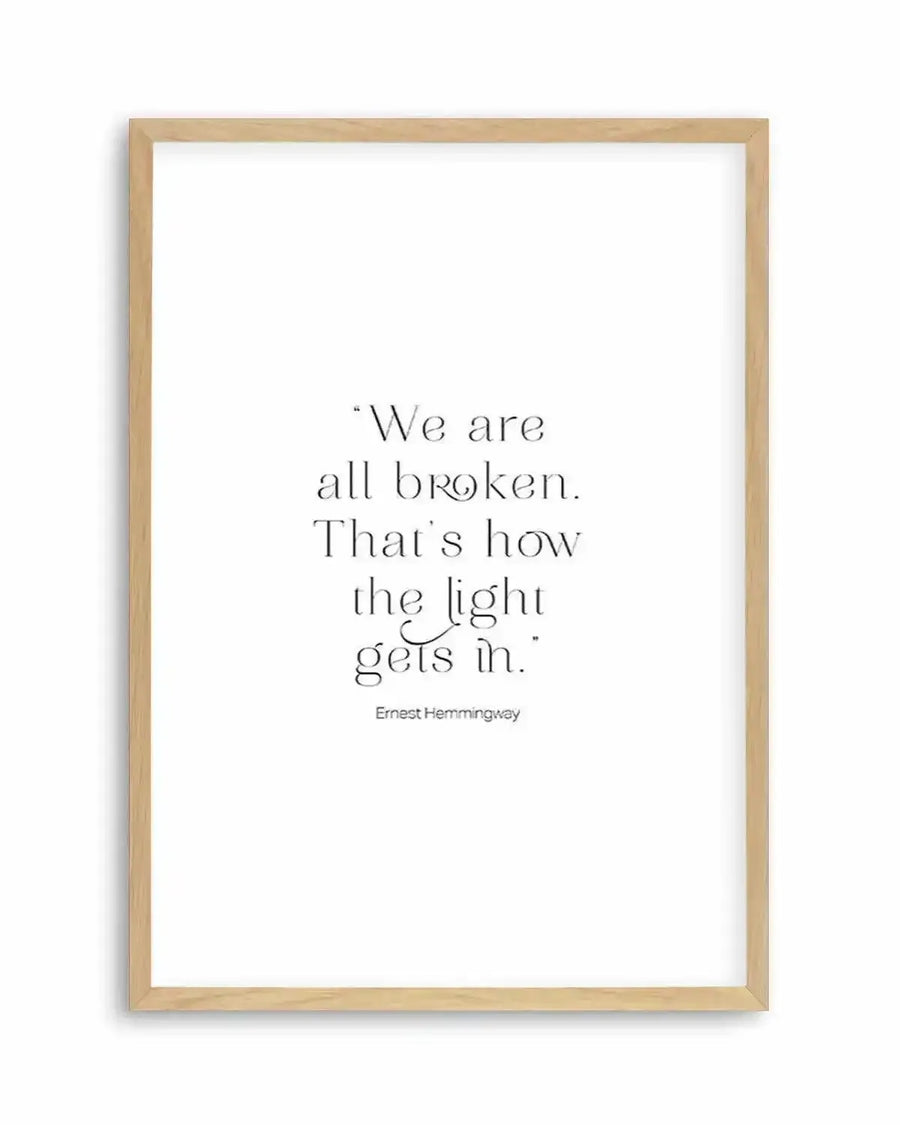 Art print featuring a quote in elegant black serif font on a crisp white background, displayed in a natural oak frame with white border. The quote reads: "We are all broken. That's how the light gets in." with "Ernest Hemingway" in smaller font below.
