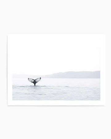 Framed black and white photograph of a whale's tail diving into calm water, with a natural wood frame and white border. The composition features the whale's flukes in the foreground, creating ripples on the water's surface, with distant mountains under a bright, overcast sky.