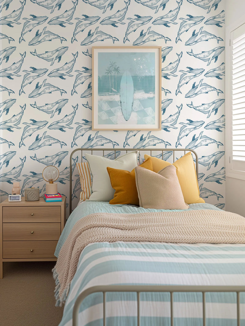 Lifestyle shot of a bedroom featuring a repeating wallpaper pattern of hand-drawn whales in navy blue on a white background. The wallpaper covers the wall behind a bed with a light grey metal frame, light blue and white striped duvet, and various pillows in cream, mustard yellow, and beige. Above the bed, a framed art print depicts a light blue surfboard on a beach with palm trees and the words "PRAY FOR SURF". A light wood nightstand with a lamp and books sits to the left of the bed.
