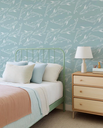 Whale Illustration in Seafoam Wallpaper featuring a repeating pattern of white line-drawn whales swimming in various directions on a soft seafoam blue background, displayed on the wall of a child's bedroom. The room includes a light green metal bed frame with white, light blue, and peach bedding, and a light wood bedside table with a white lamp.