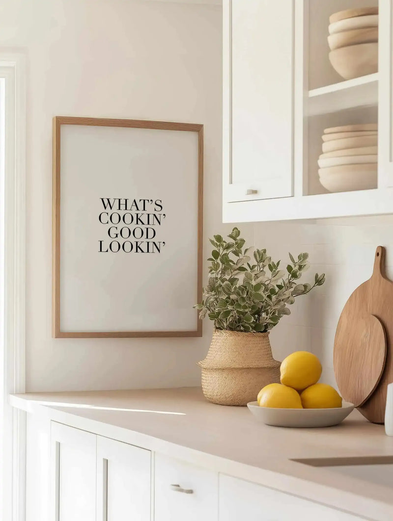 Framed art print with natural wood frame displaying "WHAT'S COOKIN' GOOD LOOKIN'" in black serif typography on cream background, hung on white kitchen wall above light wood countertop. Styled with woven basket containing green eucalyptus branches, ceramic bowl of yellow lemons, and wooden cutting board. White cabinetry with glass doors storing stacked beige bowls visible above. Minimalist, warm neutral aesthetic kitchen setting.