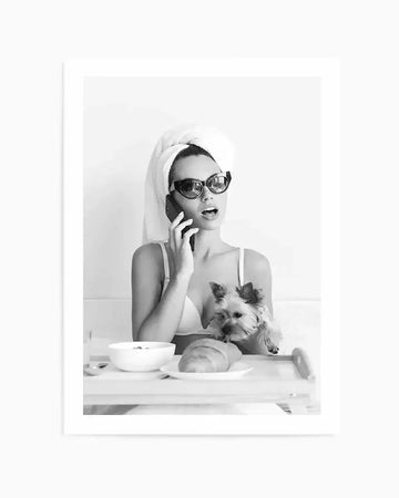Framed black and white photograph of a woman wearing a towel on her head and sunglasses, talking on a phone while having breakfast in bed with a small dog. The composition shows the woman looking surprised with her mouth open, holding a phone to her ear, with a small dog resting on the breakfast tray next to a croissant and a bowl.