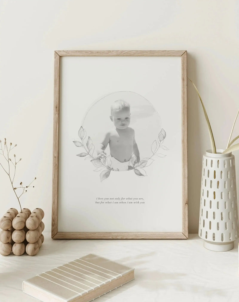 Framed black and white art print of a baby with a custom photo design, displayed in a natural oak frame with white border. The print features a baby in a circular frame with delicate leaf illustrations, and a quote below that reads "i love you not only for what you are, but for what i am when i am with you". The print is placed on a white surface next to a wooden decorative object and a white vase with dried plant stems.