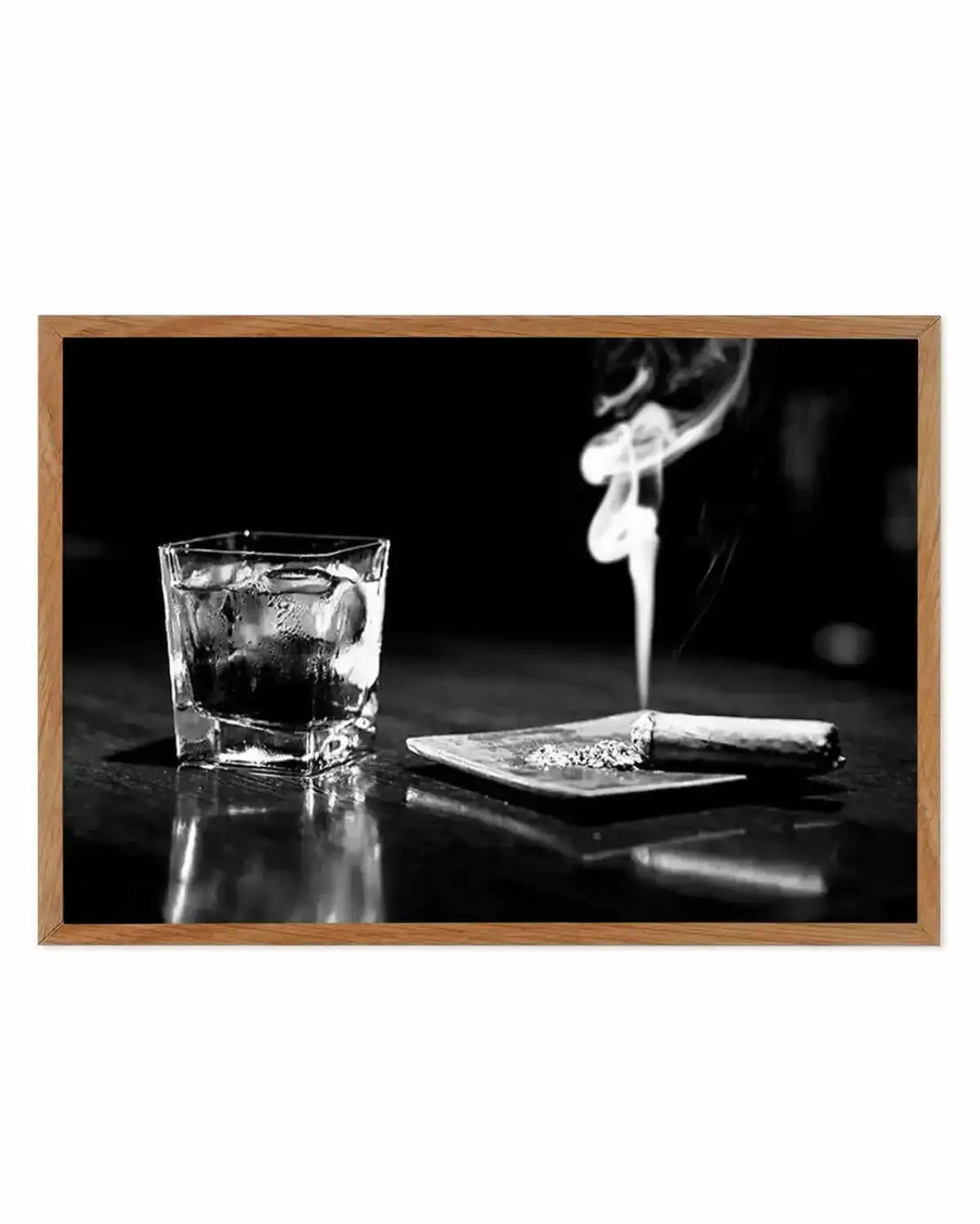 Black and white photograph showing a glass tumbler filled with dark liquor and ice on the left, a lit cigar resting on a square plate in the center with smoke rising upward, and a partial view of a person's ear wearing a dangling earring on the right. The composition is set against a dark background with reflective surface below. The image is framed in a natural wood-tone frame with no border.