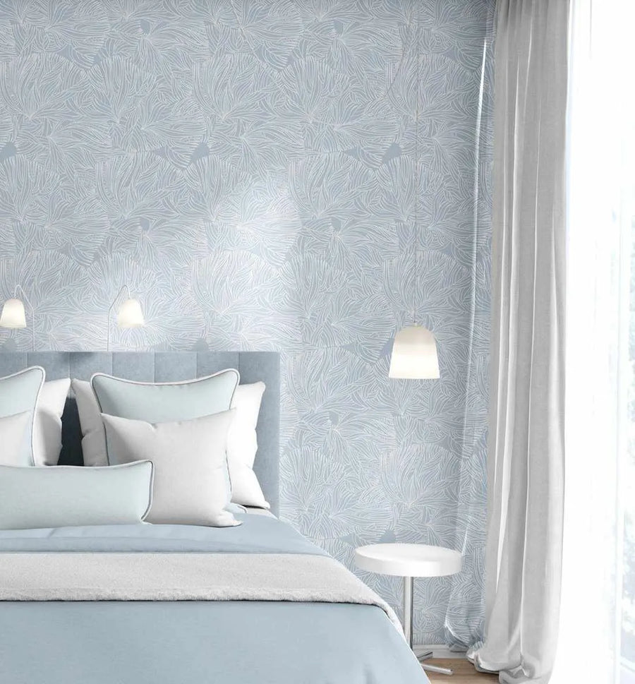Lifestyle shot of a bedroom featuring White Coral Wallpaper. The wallpaper has a repeating pattern of intricate, branching coral-like forms in white against a soft, pale blue background, creating a delicate, organic texture on the wall. The room includes a bed with a light blue upholstered headboard, white and light blue pillows, and a light blue duvet with a white throw. A white bedside table with a white lamp is visible on the right, and white curtains frame a window on the right side of the image.