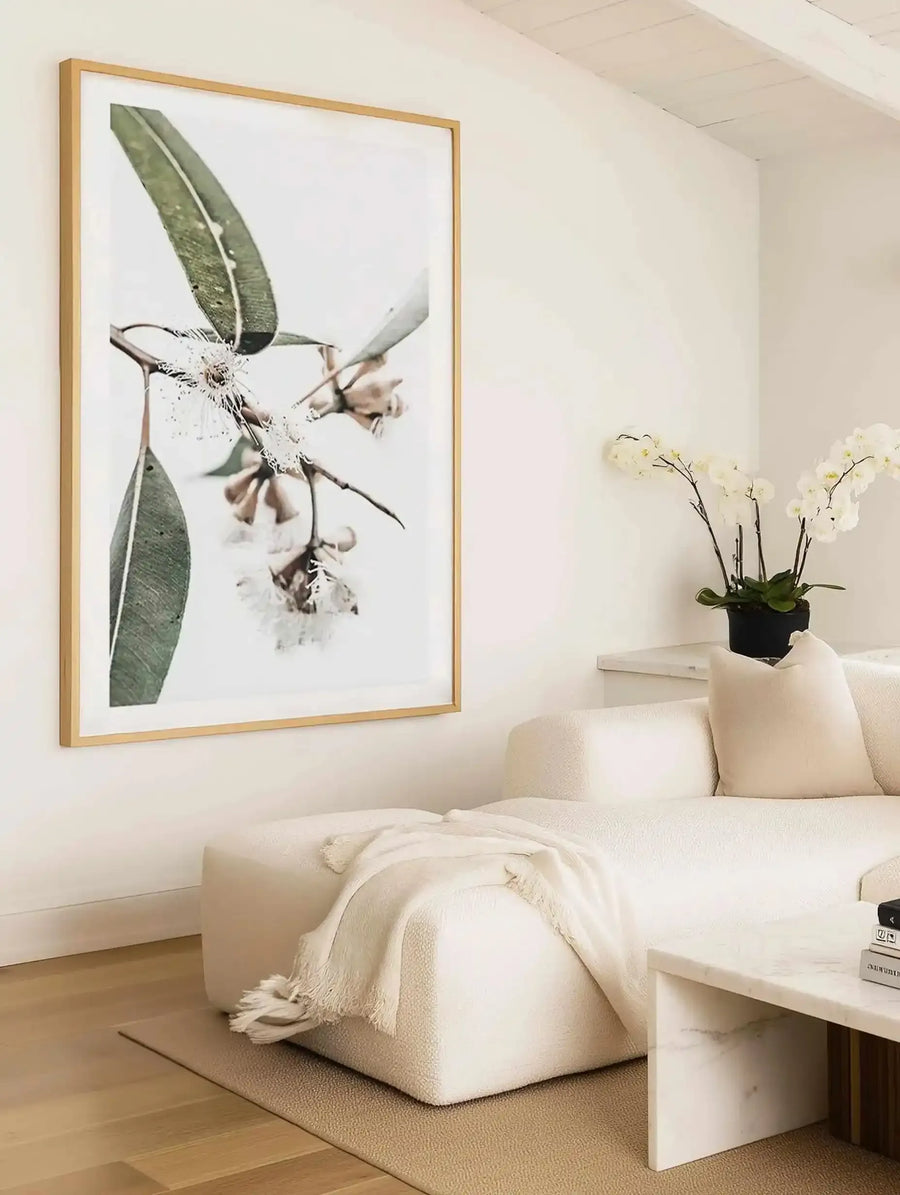Framed art print displayed above a cream-coloured curved sofa in a minimalist bedroom. The print features a botanical photograph of eucalyptus branches with silvery-green elongated leaves and delicate bell-shaped flowers in soft taupe and cream tones against a white background. The natural wood frame with white border complements the monochromatic aesthetic. A potted white orchid and cream throw blanket styled on the sofa, with light wood flooring and neutral walls completing the serene interior.