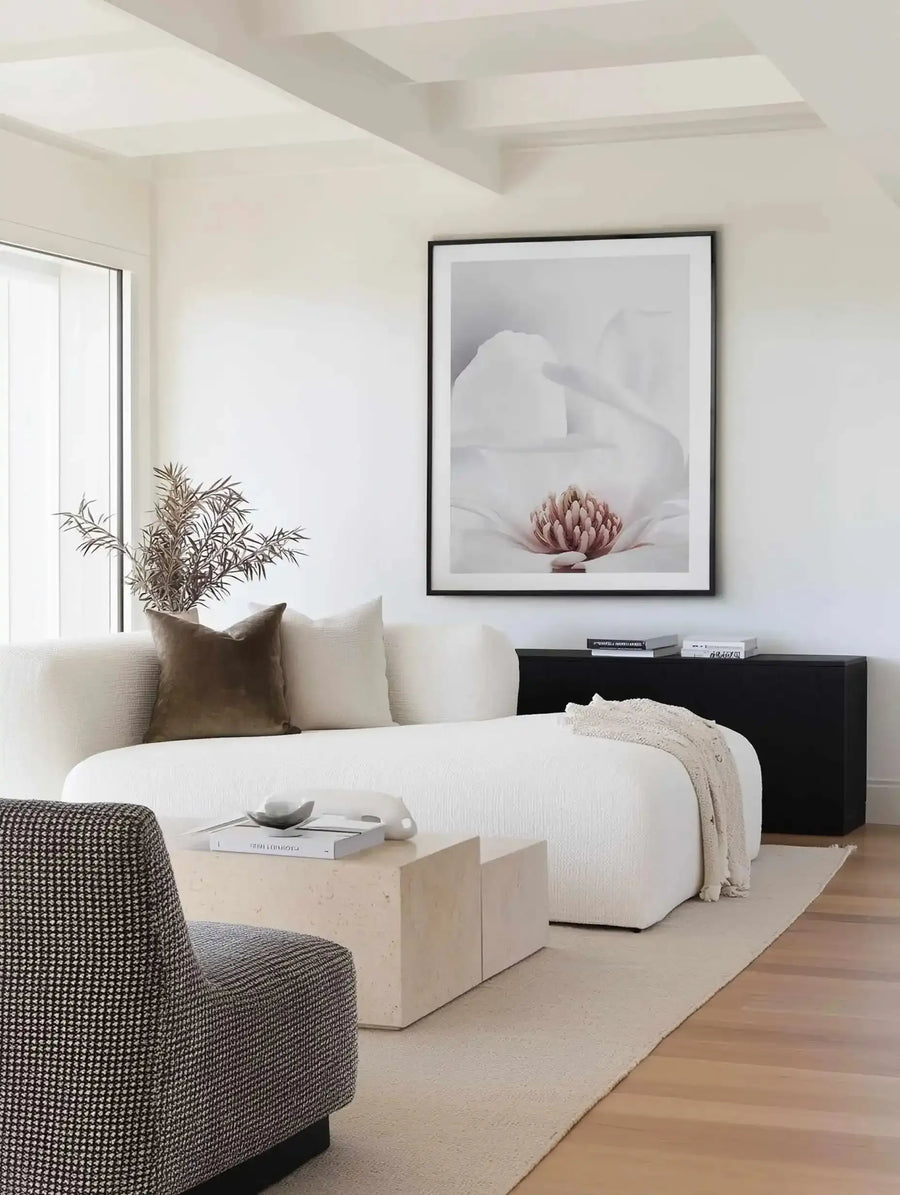 Large framed art print displayed above a black console table in a minimalist living room. The print features a close-up photograph of a white magnolia flower with soft white petals and a prominent pink and orange stamen, with a black frame and white border. The room includes a textured cream sofa with a brown velvet cushion, a cream textured throw, and dried foliage in a white vase.