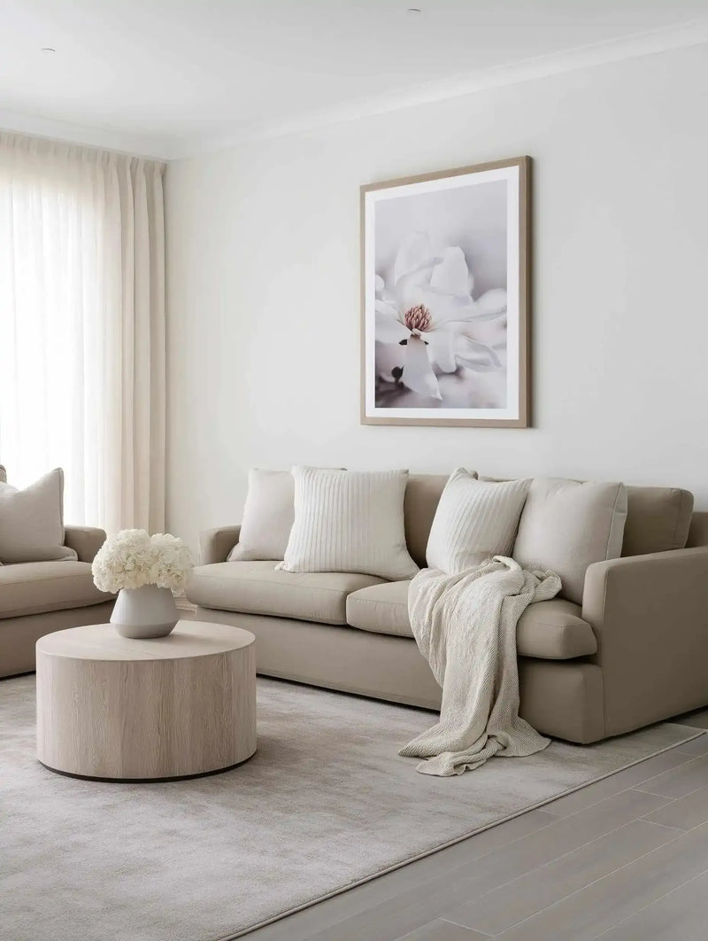 Lifestyle shot of a minimalist living room featuring a large framed art print of a white magnolia flower with a natural wood frame and white border, displayed above a beige sofa. The room includes a round light wood coffee table with a white vase of white flowers, a cream textured throw blanket, and a light grey rug.