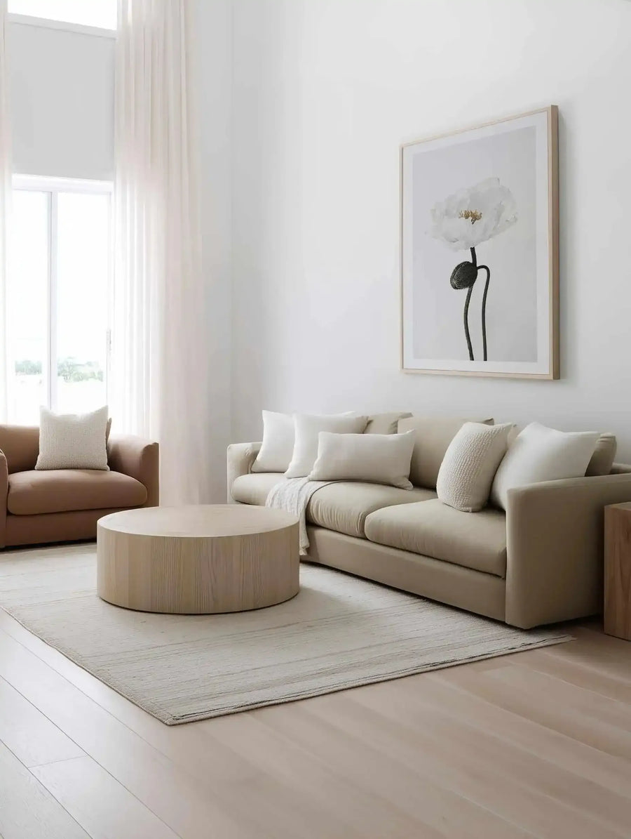 Large framed art print displayed above a neutral-toned sofa in a minimalist living room. The print features a close-up black and white photograph of a single white poppy with a dark stem and bud, set against a light grey background, with a natural wood frame and white border. The room includes a large beige sectional sofa with white throw pillows, a round light wood coffee table, a light-colored rug, and a brown armchair.
