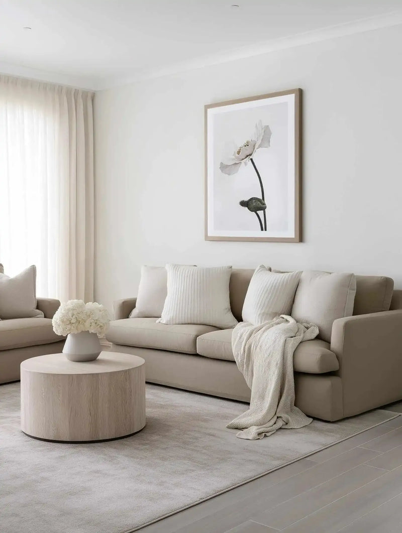 Lifestyle shot of a minimalist living room featuring a large framed art print of a single white poppy with a dark stem and bud, displayed above a light beige sofa. The print has a natural wood frame and white border. The room includes two armchairs, a round light wood coffee table with a white vase of white hydrangeas, and a textured cream throw blanket draped over the sofa.
