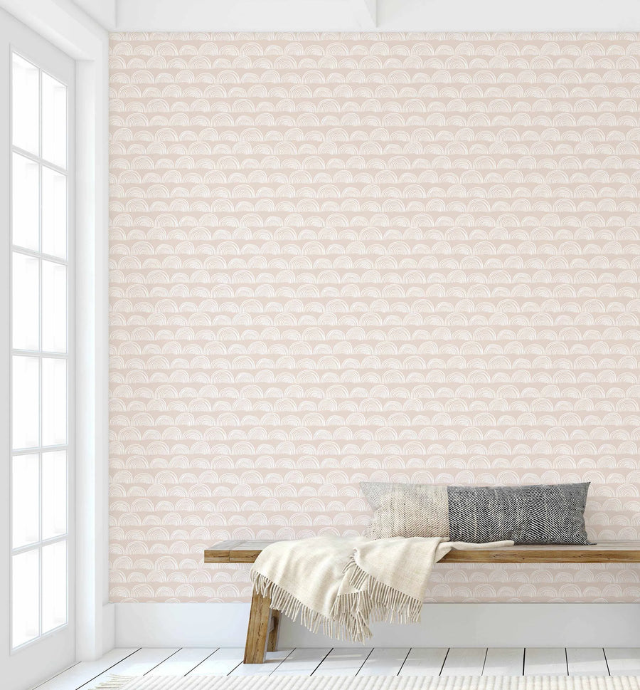 Lifestyle shot of a room with White Rainbows wallpaper. The wallpaper features a repeating pattern of hand-drawn, arching rainbow shapes in white on a soft, muted beige background. The room has white trim around a large window on the left, white wooden floorboards, and a light wood bench with a cream throw blanket and two patterned pillows (one cream and one black and white geometric) resting on it.